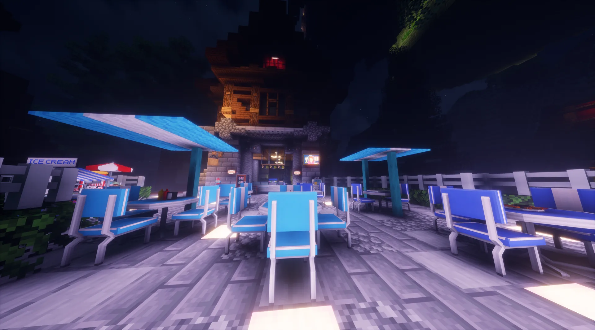 Terraza nocturna con mesas, sombrillas y tumbonas azules frente a un edificio iluminado